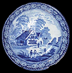 Shown on a 5 inch saucer. Men converse in front of a timbered, thatched house that has an outside bench and, possibly, an inn sign projecting from the gable on the right. The fence beyond the house suggests that this is a public building, probably an inn. The border is made up of repeated roses. An unrecorded pattern by an unknown maker, it has a TCC assigned name. Complete example from a Priv. Coll.
